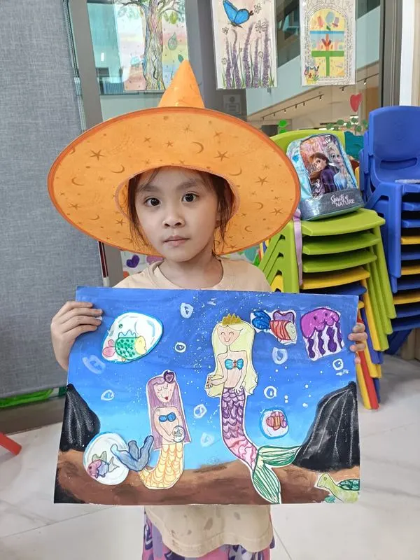 Girl wearing a fun wizard hat and proudly holding her underwater mermaid painting with jellyfish and sea creatures at Art Journey Singapore workshop