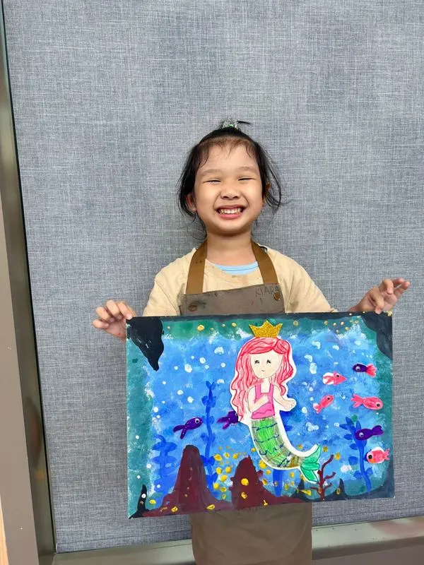 Happy child proudly holding her colourful mermaid painting completed at Art Journey Singapore