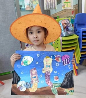 Young artist wearing a costume hat showing her underwater mermaid painting at Art Journey kids art workshop